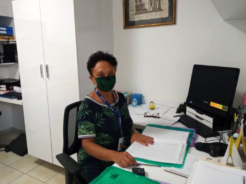 Foto da psicóloga Sueli Santos. Ela está sentada em uma cadeira de frente para uma mesa com computador, pasta e papéis. Ela usa máscara e está olhando de frente. Atrás dela há um armário branco e algumas prateleiras